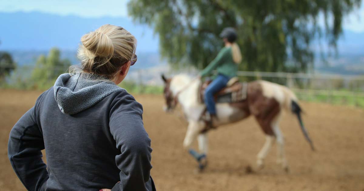 Riding instructor watching a helmeted rider on horseback in an outdoor arena