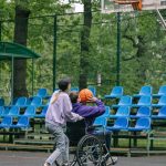 Teen in a wheelchair preparing to shoot a basketball on an outdoor court with a peer alongside