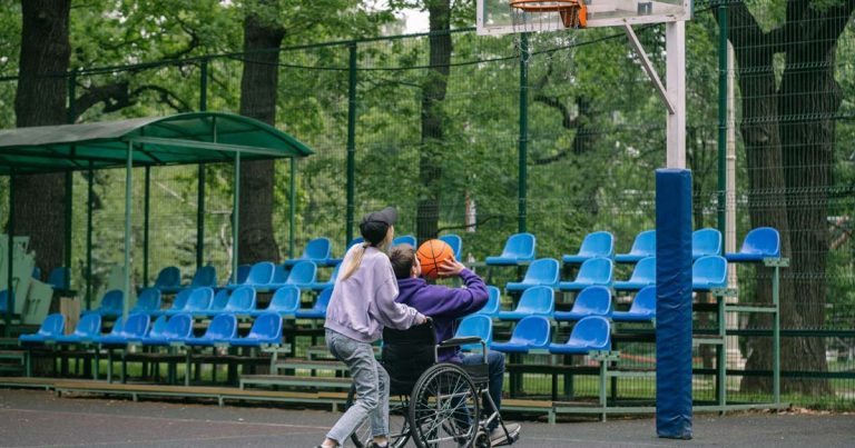 Teen in a wheelchair preparing to shoot a basketball on an outdoor court with a peer alongside
