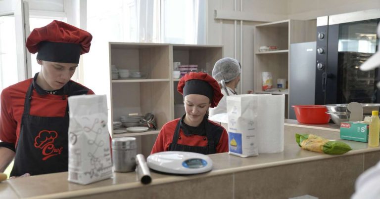 Two young adults in chef hats and aprons working side by side at a kitchen prep counter