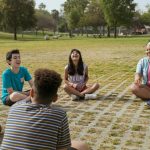 A diverse group of children and a counselor sitting in a circle outdoors at summer camp, laughing and engaged in a group activity