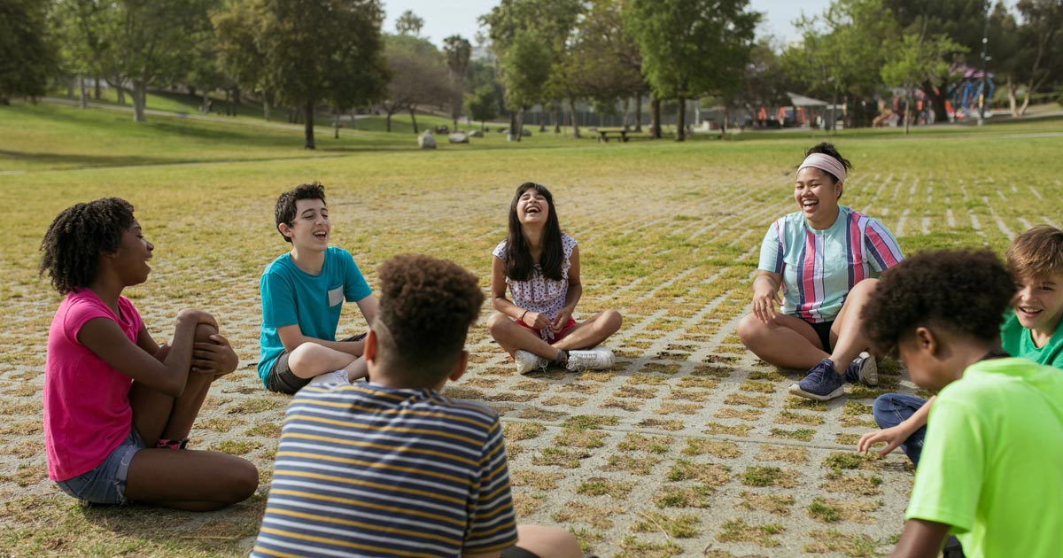 A diverse group of children and a counselor sitting in a circle outdoors at summer camp, laughing and engaged in a group activity