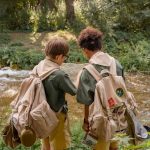 Two children with backpacks reading a map together at a stream in a wooded camp setting