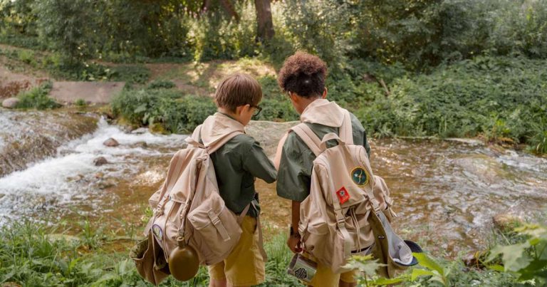 Two children with backpacks reading a map together at a stream in a wooded camp setting
