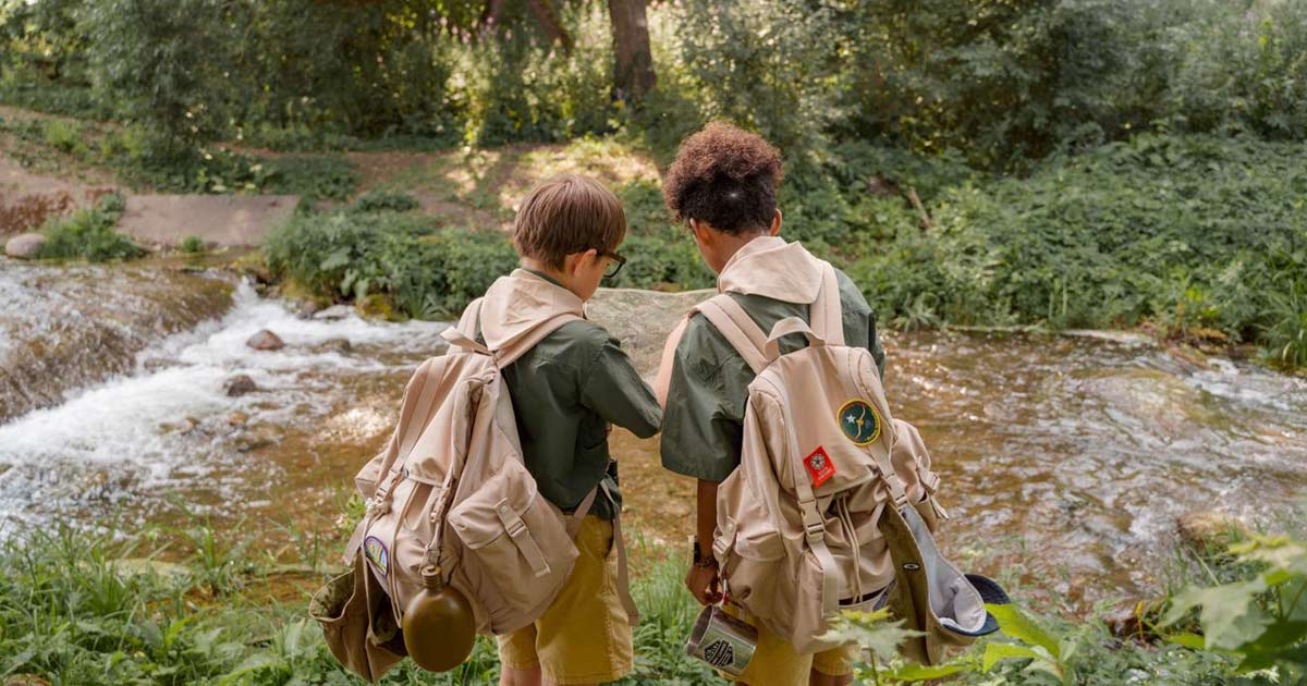 Two children with backpacks reading a map together at a stream in a wooded camp setting