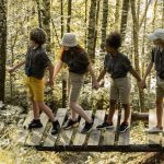 Four children crossing a wooden footbridge together through a birch forest at camp
