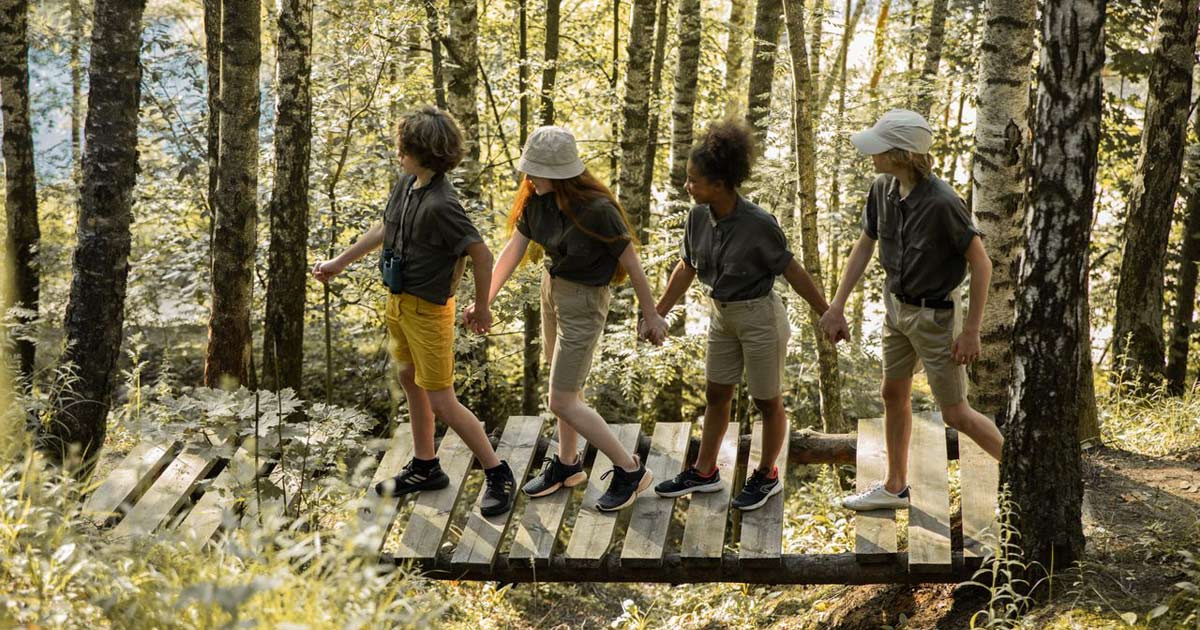 Four children crossing a wooden footbridge together through a birch forest at camp