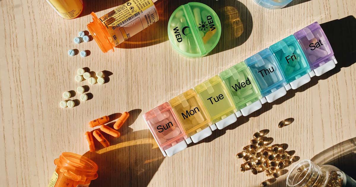 Prescription bottles and a labeled weekly pill organizer arranged on a wooden table in morning light