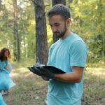 Young adult staff member writing on a clipboard while a group works along a wooded trail behind him