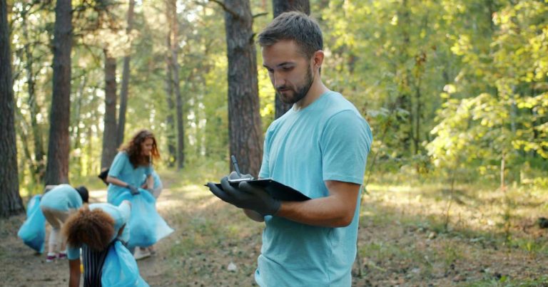 Young adult staff member writing on a clipboard while a group works along a wooded trail behind him