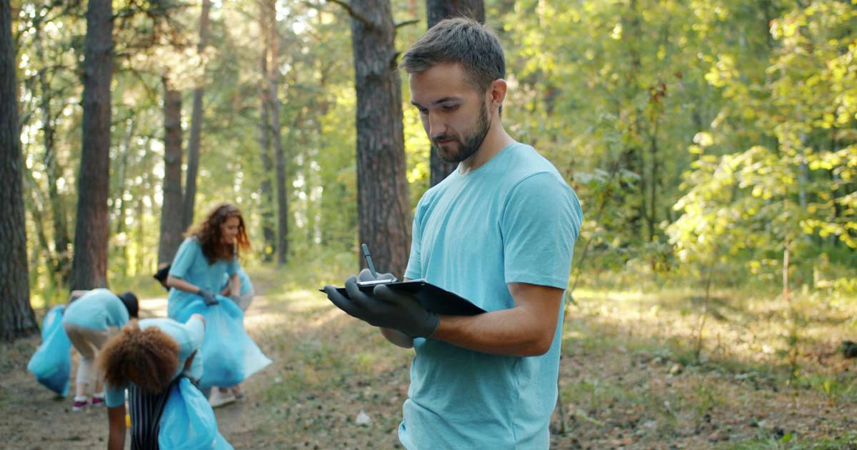 Young adult staff member writing on a clipboard while a group works along a wooded trail behind him