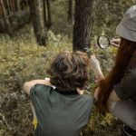 Camp staff member crouching alongside a child to examine plants with a magnifying glass outdoors