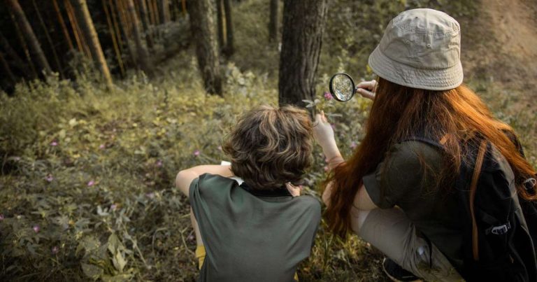 Camp staff member crouching alongside a child to examine plants with a magnifying glass outdoors