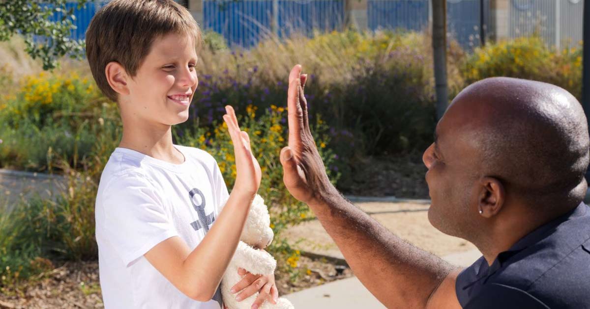 Staff member kneeling to give a high-five to a child with a stuffed animal outdoors