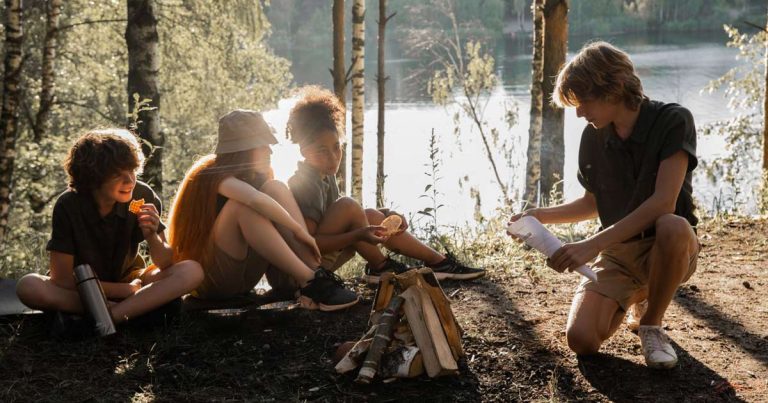 Group of teens gathered around a campfire by a lake at summer camp