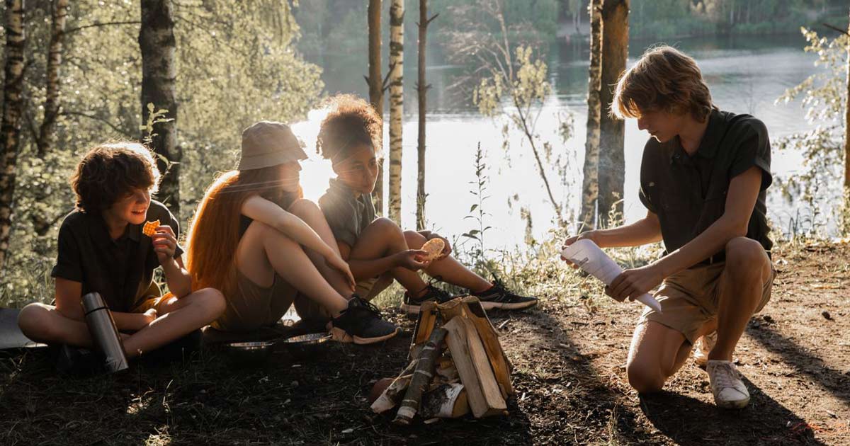 Group of teens gathered around a campfire by a lake at summer camp