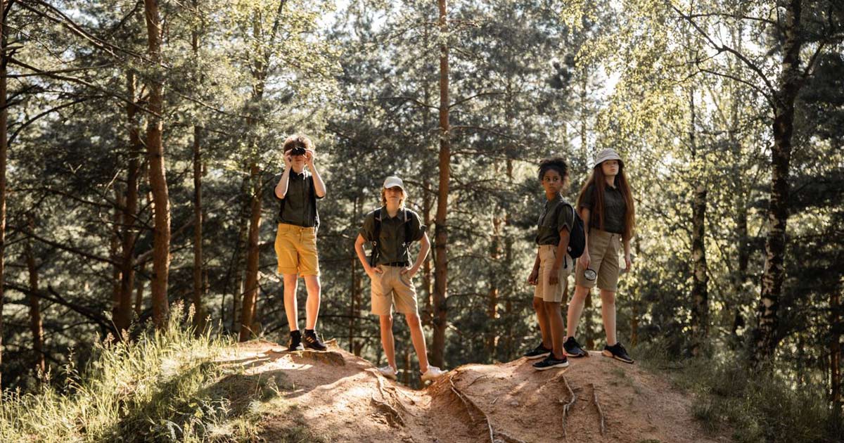 Four children in camp uniforms standing on a rocky outcrop in a pine forest