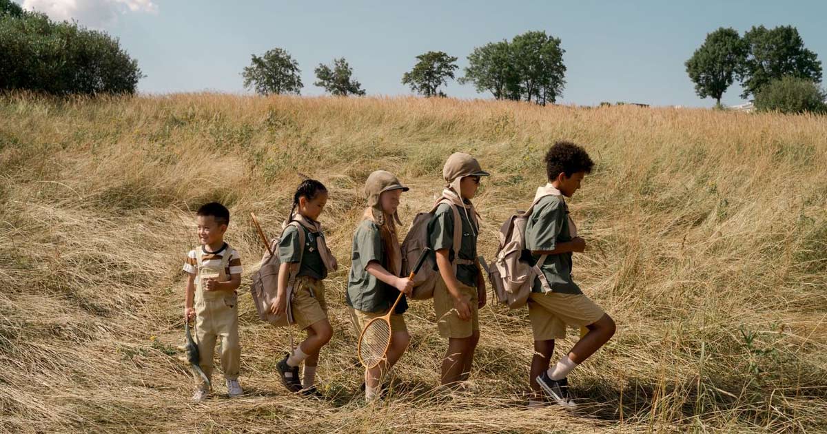 Five children in camp gear walking together through a meadow on a sunny day