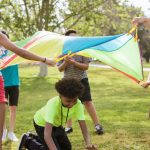 Campers and a counselor playing a cooperative parachute game together on a grassy field at summer camp