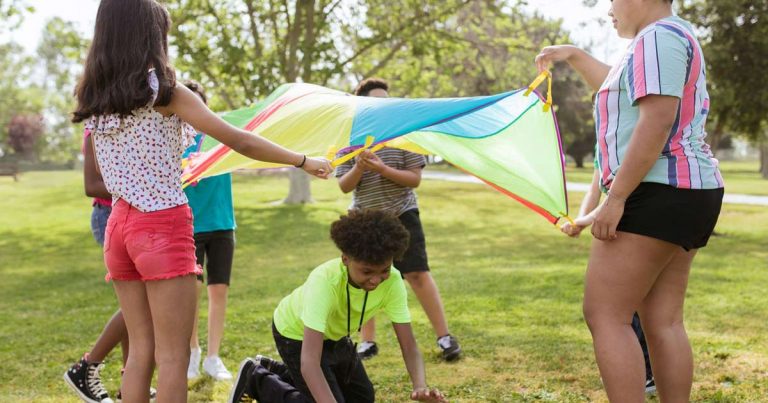 Campers and a counselor playing a cooperative parachute game together on a grassy field at summer camp