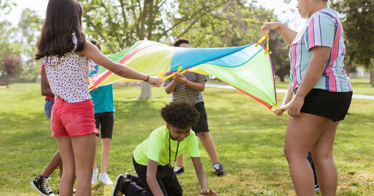 Campers and a counselor playing a cooperative parachute game together on a grassy field at summer camp