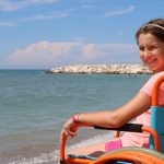 Girl smiling while sitting in a beach wheelchair at the waterfront on a sunny summer day