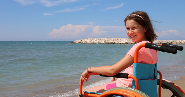 Girl smiling while sitting in a beach wheelchair at the waterfront on a sunny summer day