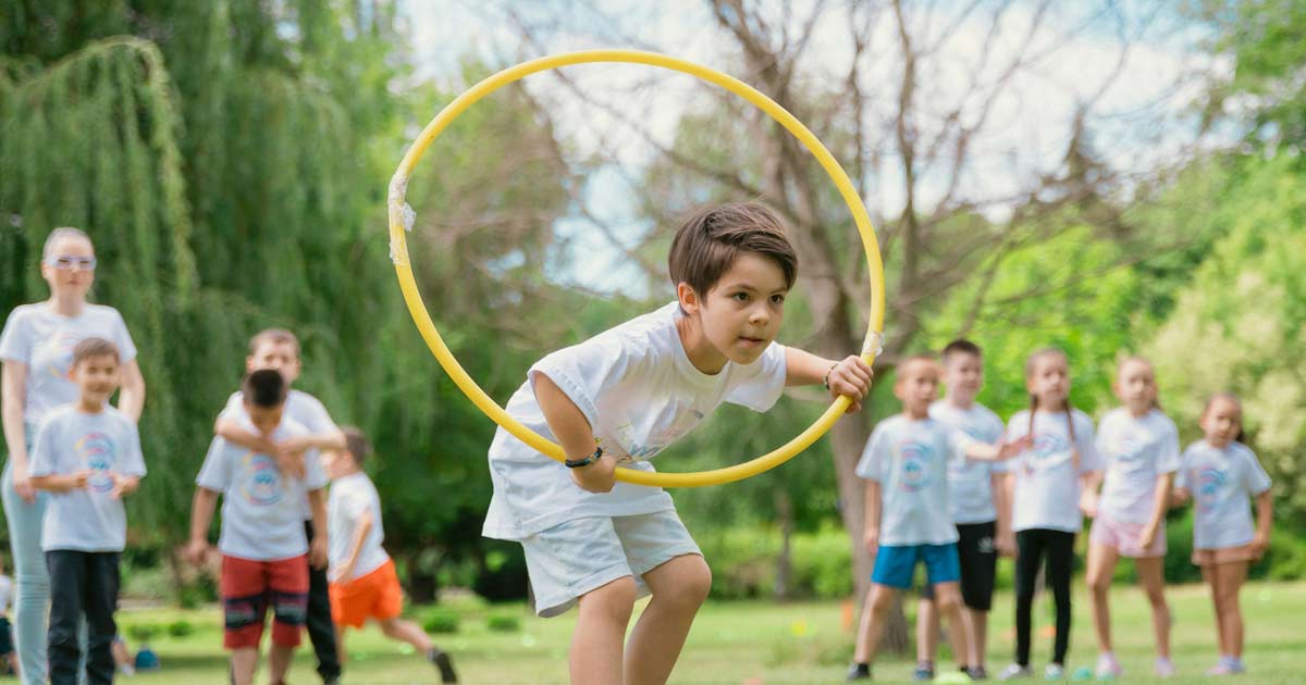 Child moving through a yellow hoop during an outdoor group activity with other participants waiting nearb